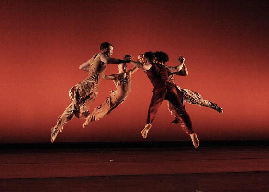 Four dancers are caught mid-air in a jump on a stage that is lit in rich, deep reds. 